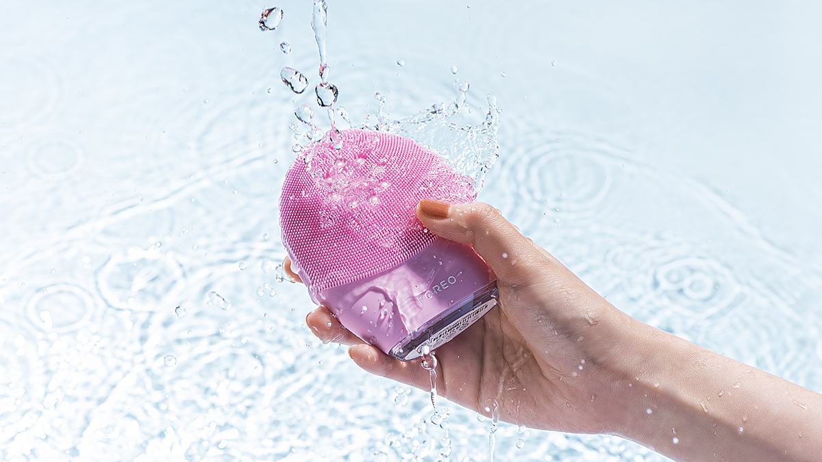 Hand holding FOREO LUNA 4 plus pink facial cleansing brush, surrounded by water