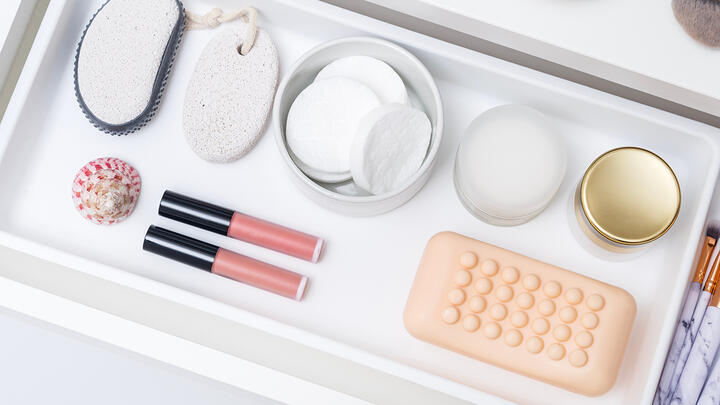 A cosmetic drawer with neatly organized soap, creams, cotton pads, pumice stone, pink lip gloss, and a sea shell