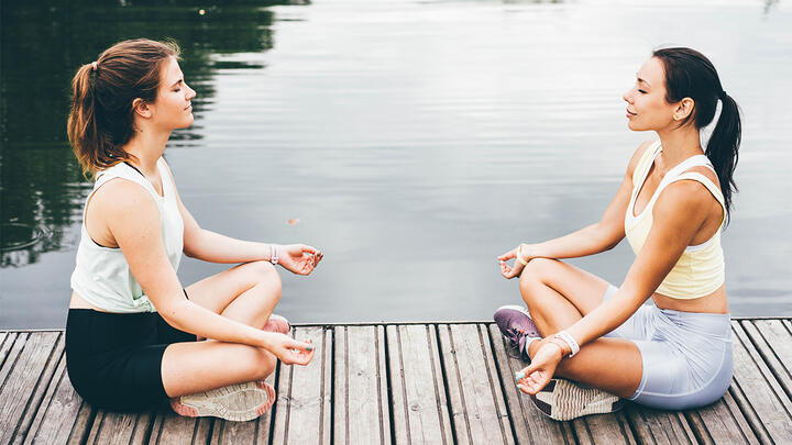 A photo of two young woman sitting on a boardwalk by the lake, meditating